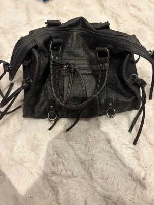 Svart handväska med flätade handtag - Säljer en svart handväska i skinn med flätade handtag och coola metalldetaljer. Väskan har dragkedja upptill, flera dekorativa remmar och ringar samt en praktisk ytterficka med dragkedja. Perfekt storlek för allt du behöver ha med dig. Väskan är ifrån Bershka.