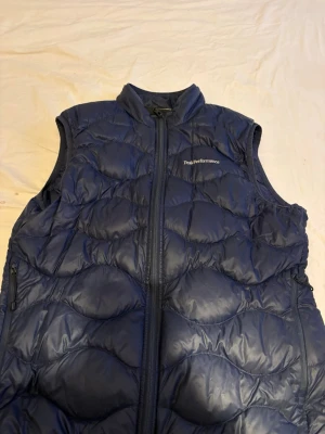 Blå dunväst från Peak Performance - Snygg blå dunväst från Peak Performance med hög krage och dragkedja framtill. Västen har två sidofickor med dragkedja och quiltad design som ger en sportig look. Perfekt att slänga över en hoodie eller tröja för extra värme.