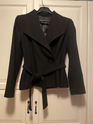 Svart kavaj med bälte från H&M - Svart kavaj från H&M med snyggt knytbälte i midjan och bred krage. Kavajen en knapp framtill och är tillverkad i en mix av viskos och ull. 