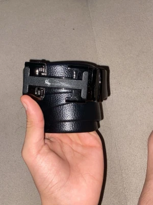 Svart bälte med H-spänne  - Säljer ett svart bälte från med ett stort H-format metallspänne. Bältet är gjort i präglat syntetmaterial som liknar skinn och har en stilren, modern look. Perfekt accessoar för att lyfta din outfit.