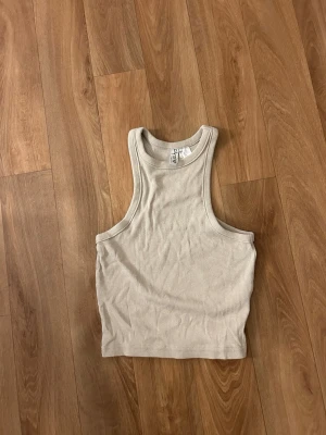 Beige ribbad linnetopp från H&M - Säljer en beige ribbad linnetopp från H&M Divided. Toppen är ärmlös med rund halsringning och croppad passform. Perfekt för varma dagar eller som lager under skjortor och jackor. Enkel och stilren design som passar till det mesta. Knappast använd
