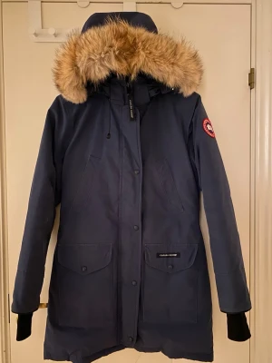 Mörkblå parkas Canada Goose Dam - Säljer en mörkblå Canada Goose parkas med stor huva och beige pälsdetalj som är avtagbar. Jackan har flera fickor framtill, dragkedja och knappar, samt den klassiska loggan på ärmen. Perfekt för kalla vinterdagar och riktigt snygg passform. Jackan har ej använts på över 10+ år och är därför i bra skick men tyget har fått lite missfärgning (se bild för detaljer). Jackan är kemtvättad. Pris kan diskuteras!