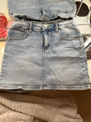 Ljusblå jeanskjol med fickor - Säljer en klassisk ljusblå jeanskjol med fem fickor och bälteshällor. Kjolen har en rak passform och stängs med knapp och dragkedja. Storlek 158 men skulle säga att den är 164. Pris går att diskutera 