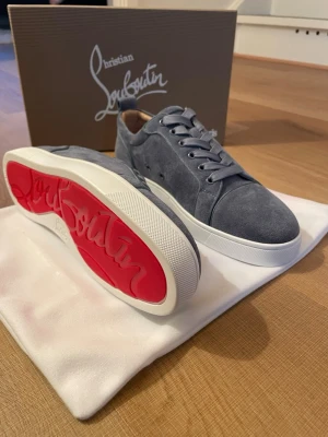 Grå mocka sneakers från Christian Louboutin - Snygga grå sneakers i mocka från Christian Louboutin med klassisk röd sula och vit kant. Skorna har rund tå, platt sula och snörning. Insidan är beige och yttersulan har Louboutin-loggan i rött. Perfekt för dig som gillar exklusiva detaljer och stilren design.