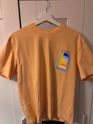 CasaBlanca peach t-shirt  - Snygg persikofärgad t-shirt CasaBlanca med tryck  fram och bak. Klassisk passform och rund hals. Storleken passar S, sitter lite oversized.