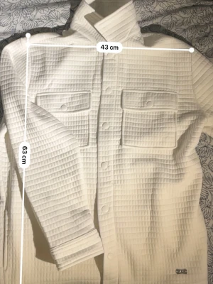 Vit quiltad overshirt med fickor - Säljer en vit overshirt med quiltad struktur och två stora bröstfickor med lock. Jackan har tryckknappar framtill och på ärmsluten, samt en klassisk krage. Perfekt för lager på lager och ger en clean look.