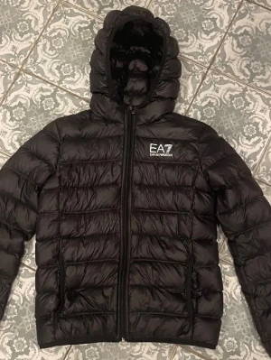 Jacka Emporio Armani - Säljer en svart pufferjacka från EA7 Emporio Armani med huva och dragkedja framtill. Jackan har ett stilrent, quiltat mönster och logga på bröstet. Perfekt för kalla dagar och riktigt snygg till streetwear-looken.