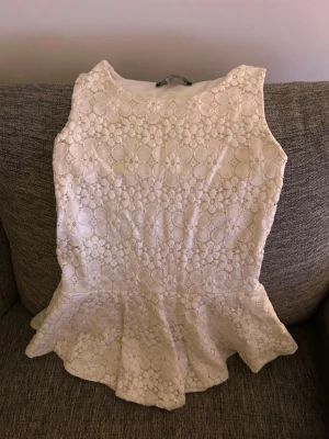 Vit spetstopp med volang - Supersöt vit topp i spets med stora blommönster och volang nertill. Ärmlös modell som ger en fräsch och somrig känsla. Perfekt att matcha med jeans eller kjol för en romantisk look.