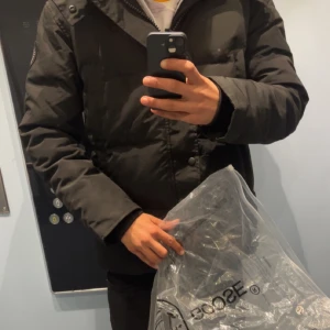 Svart pufferjacka från Canada Goose - Säljer en svart pufferjacka från Canada Goose med huva och snygga detaljer. Jackan har lång ärm, dragkedja och knappar framtill samt märkets patch på ärmen. Perfekt för kalla dagar och har en clean, modern look. Päls och påse kommer med!