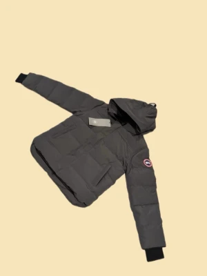 Mörkgrå dunjacka från Canada Goose - Säljer en mörkgrå dunjacka från Canada Goose med huva och klassisk logga på ärmen. Jackan har dragkedja, stora fickor framtill och ribbade muddar vid ärmsluten. Perfekt för kalla dagar och riktigt snygg till vinterlooken.Storlken är också lite liten i storlek så dne passar även M