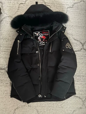 Svart dunjacka Moose Knuckles - Säljer en svart dunjacka från Moose Knuckles med fet pälskant på huvan och snygga metalldetaljer på ärmen. Jackan har dragkedjor, knappar och broderad patch på insidan. Perfekt för kalla dagar och riktigt stilren look.