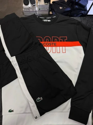 Svart och vit vindjacka från Lacoste  - Snygg vindjacka från Lacoste i svart och vitt med röd detalj och stor SPORT-logga framtill. Jackan har långa ärmar, ribbade muddar och klassisk Lacoste-logga på bröstet. Perfekt för dig som gillar sportig stil och vill sticka ut med en fräsch färgkombination.