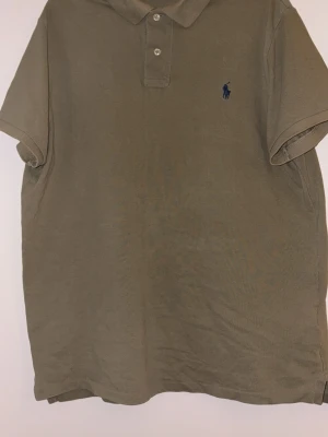  Khaki grön pikétröja från Polo Ralph Lauren - Säljer en beige pikétröja från Polo Ralph Lauren i modellen Custom Slim Fit. Tröjan har klassisk krage, två knappar framtill och den ikoniska blå broderade loggan på bröstet. Tillverkad i bomull och har korta ärmar.