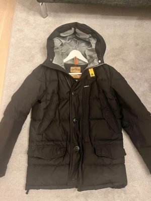 Svart dunjacka från Parajumpers XS Marcus  - Säljer en svart dunjacka från Parajumpers i storlek XS. Modell Marcus Jackan har huva, flera stora fickor, dragkedja och knappar framtill samt snygga detaljer som gul rem vid halsen. Insidan är ljusgrå och jackan är perfekt för kalla dagar. Passform slim fit. Nästintill nyskick