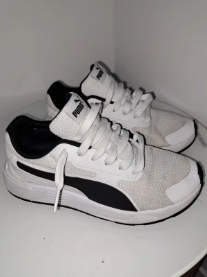 Vita och svarta sneakers från Puma - Säljer ett par vita och svarta Puma sneakers med klassisk logga på tungan och hälen. Skorna har mesh-ovandel för bra andningsförmåga, snörning och en bekväm platt sula. Perfekta för dig som gillar sportig och clean stil. Size 38 passar 39 och även 40. Lägg gärna prisförslag 
