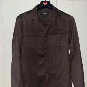Mörkbrun overshirt från Santini - Mörkbrun overshirt med klassisk krage och två bröstfickor med lock. Skjortan har långa ärmar och knäppning framtill med svarta knappar. Perfekt att slänga över en t-shirt för en chill look.
