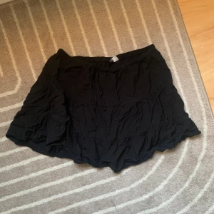 Svart volangkjol från H&M Divided - Supersöt svart kortkjol från H&M Divided med flera volanglager och resår i midjan. Kjolen är luftig och har en lätt rynkad struktur som ger en avslappnad och trendig look. Perfekt att styla med en crop top eller oversized hoodie.