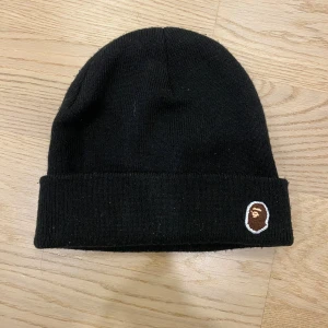 Svart mössa från A Bathing Ape - Svart ribbstickad mössa från A Bathing Ape med klassisk uppvikt kant och den ikoniska BAPE-loggan broderad framtill. Enkel och stilren design som passar perfekt till streetwear-looken.