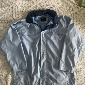 Ljusblå vindjacka från True North - Säljer en ljusblå vindjacka från True North med marinblå detaljer vid kragen. Jackan har dragkedja framtill, hög krage som går att vika ut till en huva och är tillverkad i lätt polyester. Perfekt för blåsiga dagar och enkel att slänga på sig över en hoodie. Jackan har en liten rispa på ryggen, se sista bilden 
