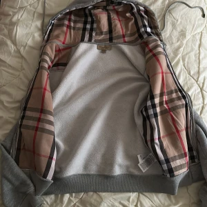 Byte elle köp - Säljer en grå hoodiejacka från Burberry i bomull med klassiskt rutigt foder i beige, svart och rött. Jackan har dragkedja, snörning i huvan och ribbade muddar. Perfekt för dig som vill ha en stilren och lyxig streetwear-look.