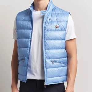 Ljusblå dunväst från Moncler - Snygg ljusblå dunväst från Moncler med quiltad design och hög krage. Västen har två dragkedjefickor framtill och klassisk Moncler-logga på bröstet. Perfekt att slänga över en t-shirt för en sportig look. FINNS I FLERA STORLEKAR 