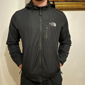 Svart vindjacka The North Face 2XL - Svart vindjacka från The North Face Summit Series med huva, dragkedja framtill och flera fickor med dragkedja. Jackan har en clean look, justerbar nederkant och logga på bröstet och ryggen. Perfekt för dig som vill ha en stilren och funktionell jacka. 