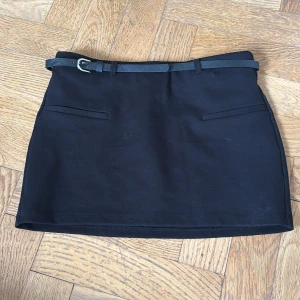 Svart kortkjol med bälte från Zara - Svart kortkjol från Zara med smalt bälte i midjan och två fickor framtill. Kjolen har en klassisk, rak passform och är tillverkad i ett mjukt material. I Storlek S