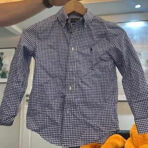 Blårutig skjorta från Ralph Lauren (barn, 130) - Snygg blårutig skjorta från Ralph Lauren med klassisk button-down krage och broderad logga på bröstet. Skjortan har lång ärm och är gjord i mjuk bomull. Perfekt för dig som gillar stilrena och tidlösa plagg med en preppy vibe.