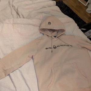 Beige hoodie från Sail Racing - Säljer en beige hoodie från Sail Racing med svart logga på bröstet och tryck på huvan. Tröjan har känguruficka, snörning i huvan och är riktigt skön och stilren. Perfekt för dig som gillar streetwear och vill ha något snyggt och bekvämt.