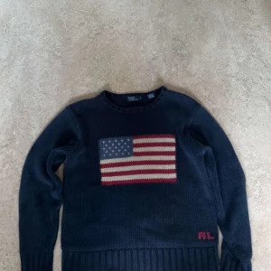 Mörkblå stickad tröja Polo Ralph Lauren - Snygg mörkblå stickad tröja från Polo Ralph Lauren i storlek S. Framsidan har en stor amerikansk flagga i rött, vitt och blått, samt RL-broderi i rött nedtill. Tröjan har rund hals och långa ärmar med ribbade muddar. Perfekt för dig som gillar klassisk och cool stil.