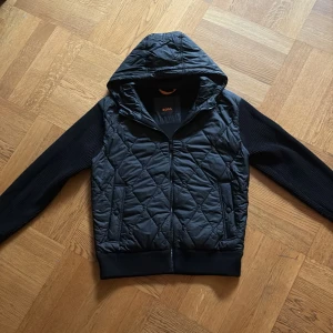 Svart quiltad cardigan jacka från BOSS - Svart cardigan från BOSS med quiltad framsida och stickade ärmar. Jackan har dragkedja, huva och ribbade muddar. Snygg BOSS-logga på ärmen och en clean, modern look. Perfekt för dig som gillar streetstyle och vill ha något som sticker ut. Skriv om det finns frågor 