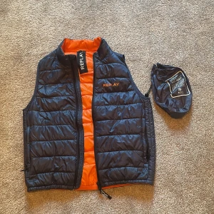 Svart dunväst från Replay med orange foder - Snygg svart dunväst från Replay med quiltad design och orange insida. Västen har dragkedja framtill, hög krage och Replay-logga på bröstet. Levereras med tillhörande packpåse, perfekt att ta med på språng. Lätt och smidig modell.