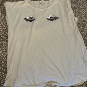 Vit ärmlös topp med paljettögon Lindex - Vit ärmlös topp från Lindex med två stora ögon framtill, dekorerade med lila och silvriga paljetter och svarta fransiga fransar som ögonfransar. Mjuk och skön bomullskänsla, perfekt för dig som gillar unika detaljer.