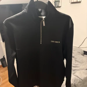 Svart half zip-tröja från Les Deux - Svart långärmad tröja från Les Deux med stilren half zip-dragkedja och hög krage. Tröjan har diskret broderad logga i guld på bröstet och är gjord i ett mjukt, slätt material som känns skönt mot huden. Perfekt för en clean och modern look.
