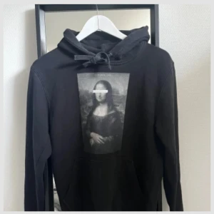 Svart hoodie Mona Lisa The Cool Elephant - Svart hoodie från The Cool Elephant med stor Mona Lisa-print på bröstet. Klassisk känguruficka, huva med snörning och mjukt fleecetyg på insidan. Perfekt för dig som gillar streetstyle och konstnärliga detaljer. Säljer pågrund av att den ej passar mig längre