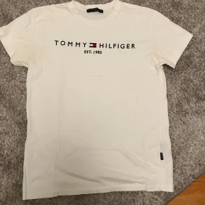 Vit t-shirt Tommy Hilfiger M - Vit t-shirt från Tommy Hilfiger med klassisk logga och texten 'EST. 1985' broderad på bröstet. T-shirten har rund hals, korta ärmar och är gjord i mjuk bomull. Perfekt basic-plagg med snygg passform och diskret logotagg i nederkant.