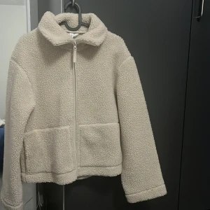 Beige teddyjacka med dragkedja - Mysig beige teddyjacka med hög krage och hel dragkedja framtill. Jackan har två stora fickor och är tillverkad i fluffigt fleece-material som håller dig varm under kyliga dagar. Perfekt för lager-på-lager och chill vardagsstil.