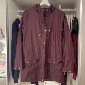 Vinröd regnjacka med huva från Cubus - Säljer en vinröd regnjacka från C i storlek XS. Jackan har huva, dragsko i halsen, tryckknappar och stora fickor framtill. Långärmad modell med slät yta och innerfoder i vitt. Perfekt för regniga dagar.