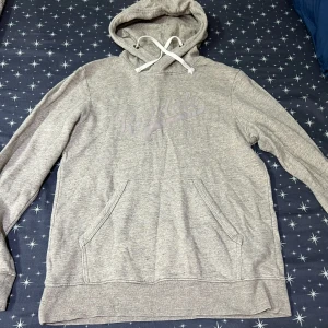 Grå hoodie från H&M med tryck - Gråmelerad hoodie från H&M med trycket 'Rexdale' framtill. Tröjan har huva med vita snören och en stor magficka. Perfekt för chill dagar och enkel att matcha med jeans eller joggers.