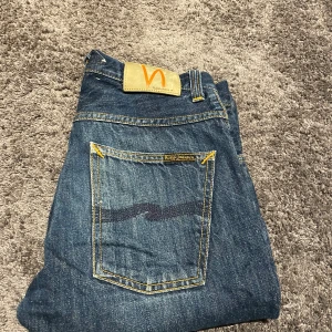 Blå Nudie Jeans 28/32 straight fit - Säljer ett par klassiska blå jeans från Nudie Jeans i storlek 28/32. Jeansen har raka ben, snygga slitningar och kontrastsömmar i gult. Bakfickorna har Nudie Jeans signaturbrodyr och patch i beige med orange logga. Perfekta för en avslappnad stil.
