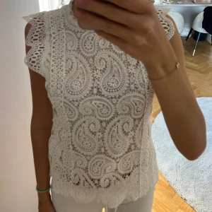Vit spetsblus med paisleymönster - Superfin vit blus i spets med detaljerat paisleymönster och små blommor. Blusen är ärmlös med volangkant vid axlarna.