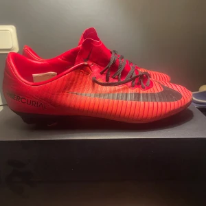 Nike Mercurial röda fotbollsskor - Säljer ett par Nike Mercurial fotbollsskor i en riktigt snygg röd färg med svarta detaljer och snörning. Skorna har en låg profil och är designade för att ge bra grepp på planen. Materialet är syntet och har ett randigt mönster som ger en sportig vibe.