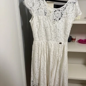 Vit spetsklänning med öppen rygg - Superfin vit klänning i spets med blommigt mönster och öppen rygg. Klänningen har korta ärmar och markerad midja, vilket ger en romantisk och somrig känsla. Perfekt för dig som gillar detaljer och vill sticka ut med en elegant look.