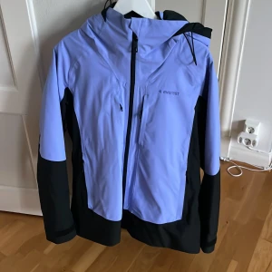 Ljuslila och svart skidjacka Everest - Skidjacka från Everest i ljuslila och svart med huva och dragkedja framtill. Jackan har flera praktiska fickor med dragkedja och justerbar huva. Ficka finns även i vänster ärm för att exempelvis förvara liftkort. Har använts fåtal gånger just vid skidåkning, vilket den har passat utmärkt till. Säljer nu pga. lite för stor för mig.