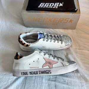 Golden goose glitter och leopard - Unika Golden Goose sneakers i vitt skinn med silverglittrig tå, rosa stjärna på sidan och leopardmönstrad häl. Grå snörning och texten 'LOVE NEVER CHANGES' på sulan. Perfekta för dig som vill sticka ut med coola detaljer och mixade material. Storlek 39 men skulle säga att de passar 38 bättre. Perfekt nu till hösten, 0bs lnte 0rglnal men mycket bra kvalitet 😊