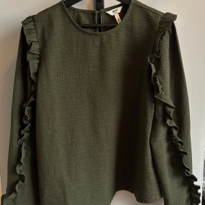 Olivgrön tröja med volang från Object - Snygg olivgrön tröja från Object med lång ärm och volangdetaljer längs axlar och ärmar. Tröjan har en lätt strukturerad yta och rund halsringning. Perfekt för dig som vill ha en stilren men ändå unik look.