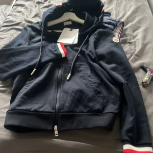 Svart Moncler bomberjacka med huva - Svart bomberjacka från Moncler med huva och dragkedja. Jackan har ribbade muddar med röd och vit detalj, samt Moncler-logga på ärmen. Snygga snören i huvan och klassisk passform. Perfekt för dig som vill ha en stilren och sportig look. Passar bra för bra på s