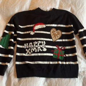Svart jultröja med ränder från H&M - Svart och vitrandig jultröja från H&M med broderade julmotiv som pepparkaka, julgran, paket och tomteluva. Texten 'Happy Xmas' framtill. Långärmad och rund hals. Perfekt för julens alla tillfällen!