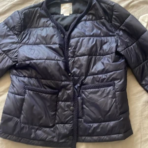 Mörkblå pufferjacka från Gina Tricot - Snygg mörkblå pufferjacka från Gina Tricot med quiltad design och stora framfickor. Jackan har tryckknappar framtill och är tillverkad i glansig polyester. Perfekt för kyliga dagar och ger en clean look.