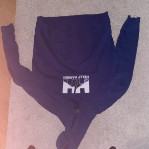 Blå Helly Hansen hoodie med tryck - Säljer en blå hoodie från Helly Hansen med stor vit logga och tryck på bröstet. Tröjan har huva med snörning och en klassisk känguruficka framtill. Perfekt för dig som gillar sportig och avslappnad stil.